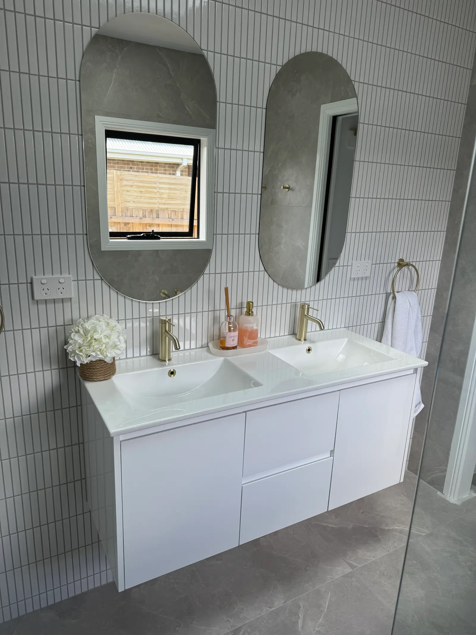 Wall-hung white double vanity with brass tapware and two oval mirrors on a white finger-tile wall