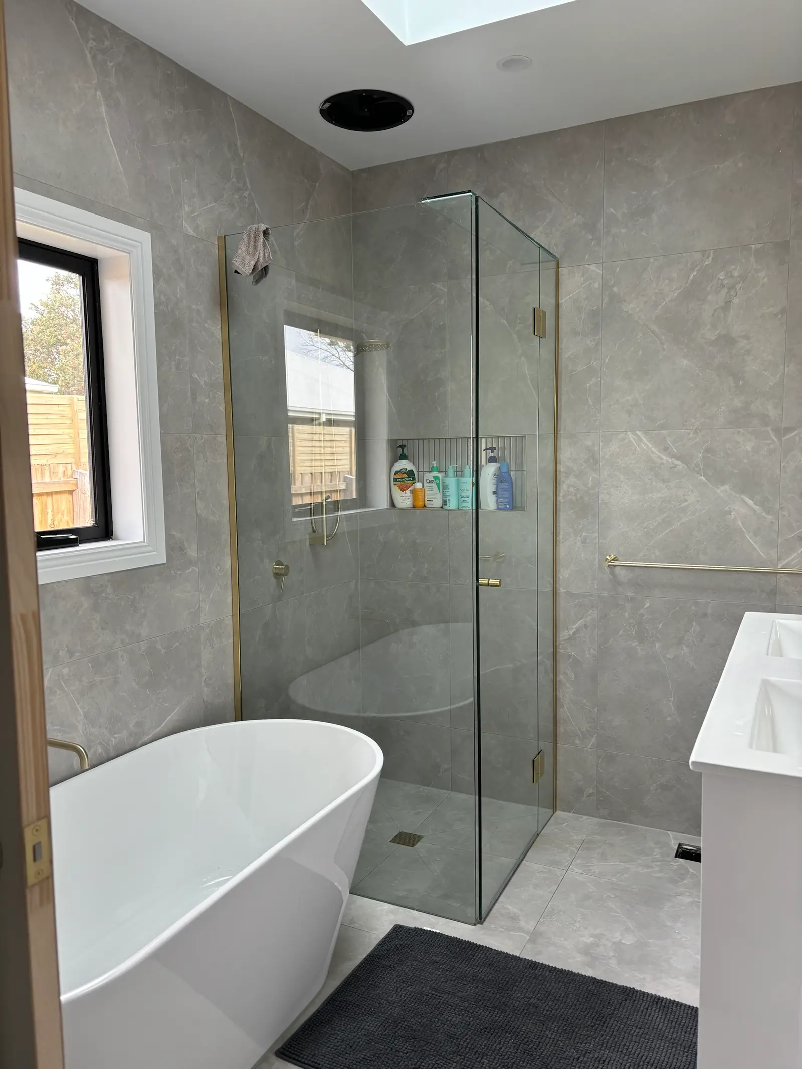 Frameless brass-trimmed shower screen beside a freestanding bath, with a tiled shower niche