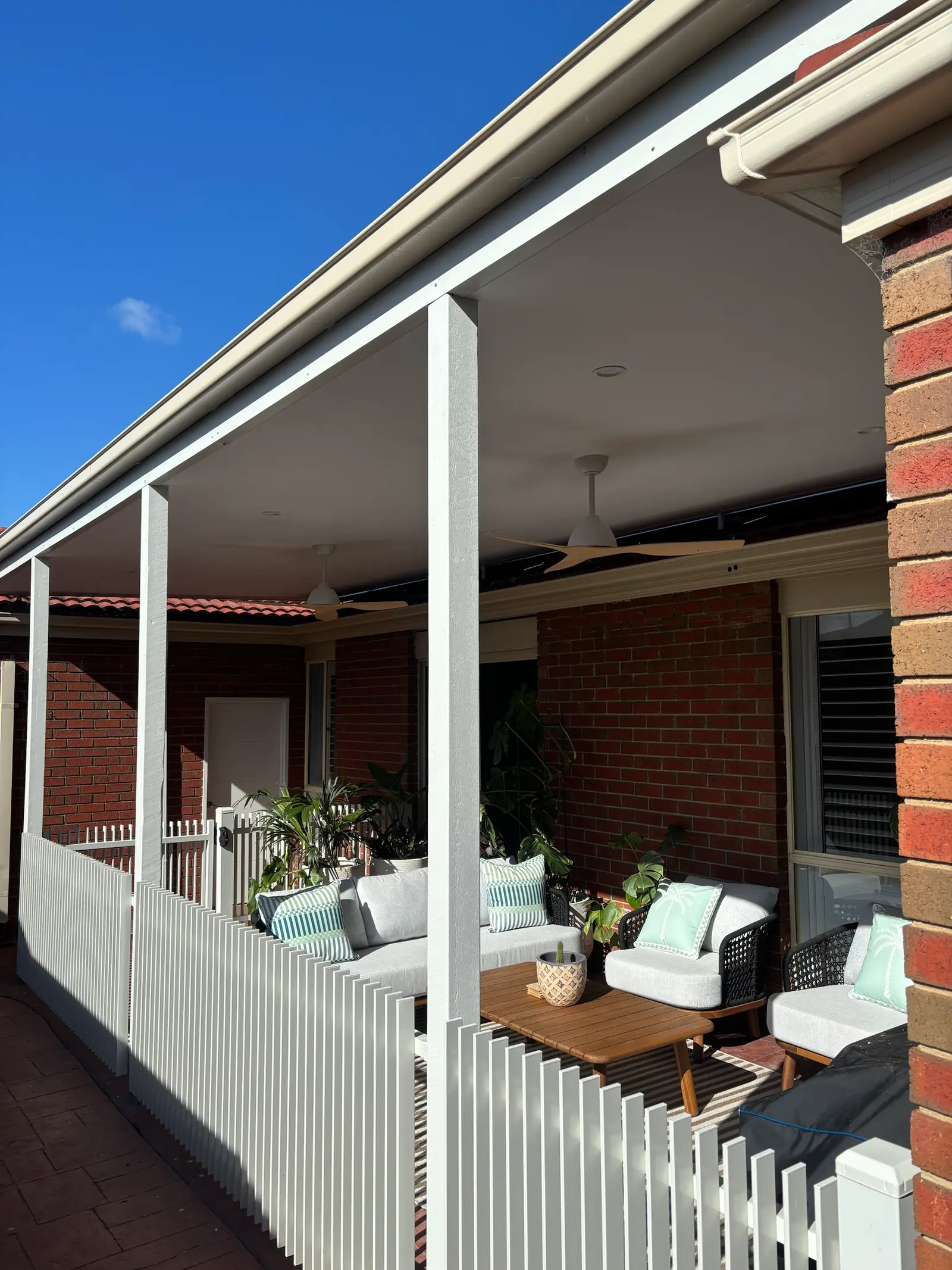 Flat-roof verandah on a red brick home with painted posts, lined ceiling, ceiling fans and outdoor seating behind a white picket fence