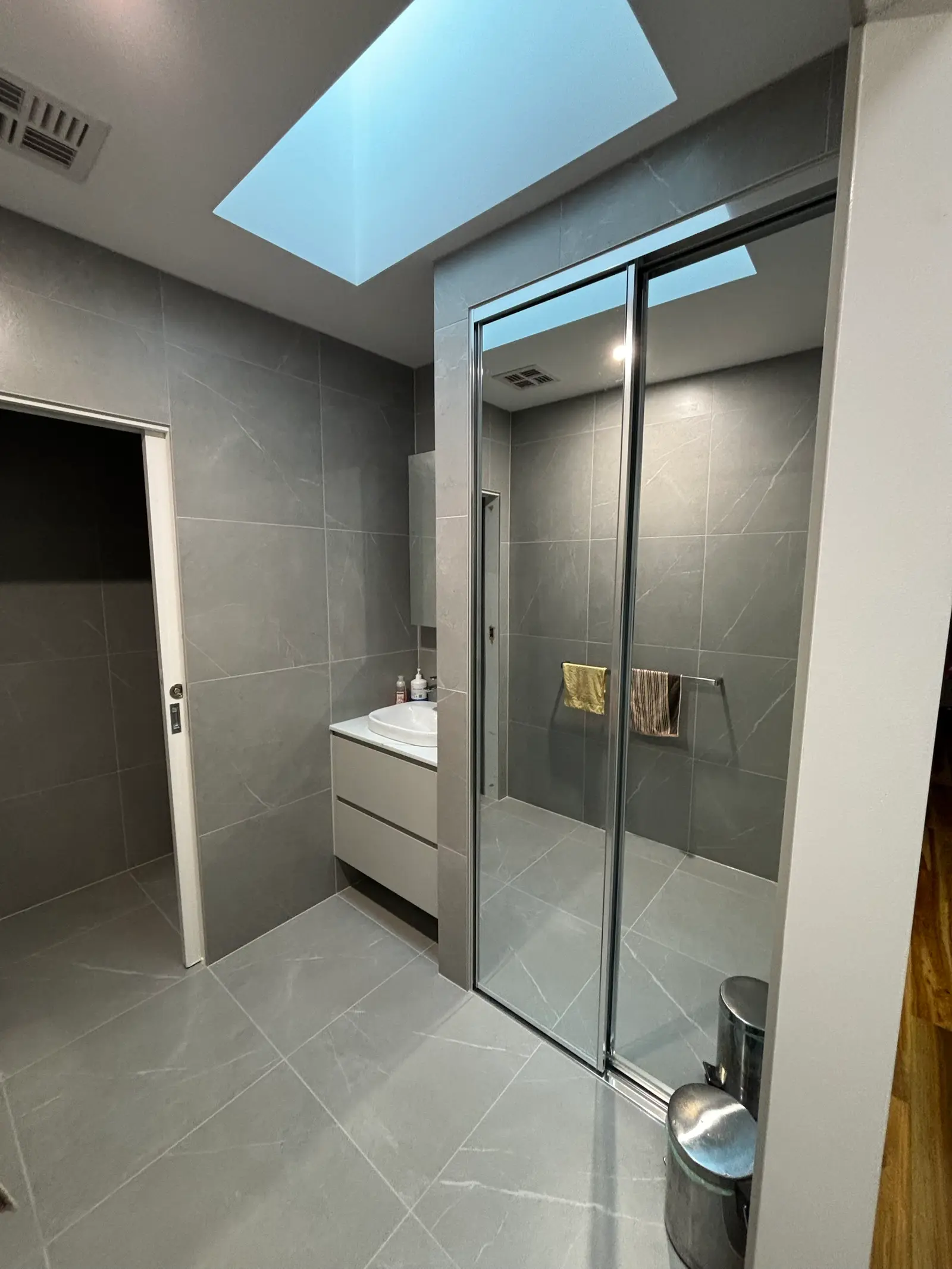 Renovated grey-tiled powder room with a skylight overhead, wall-hung vanity with single basin, and mirrored sliding doors