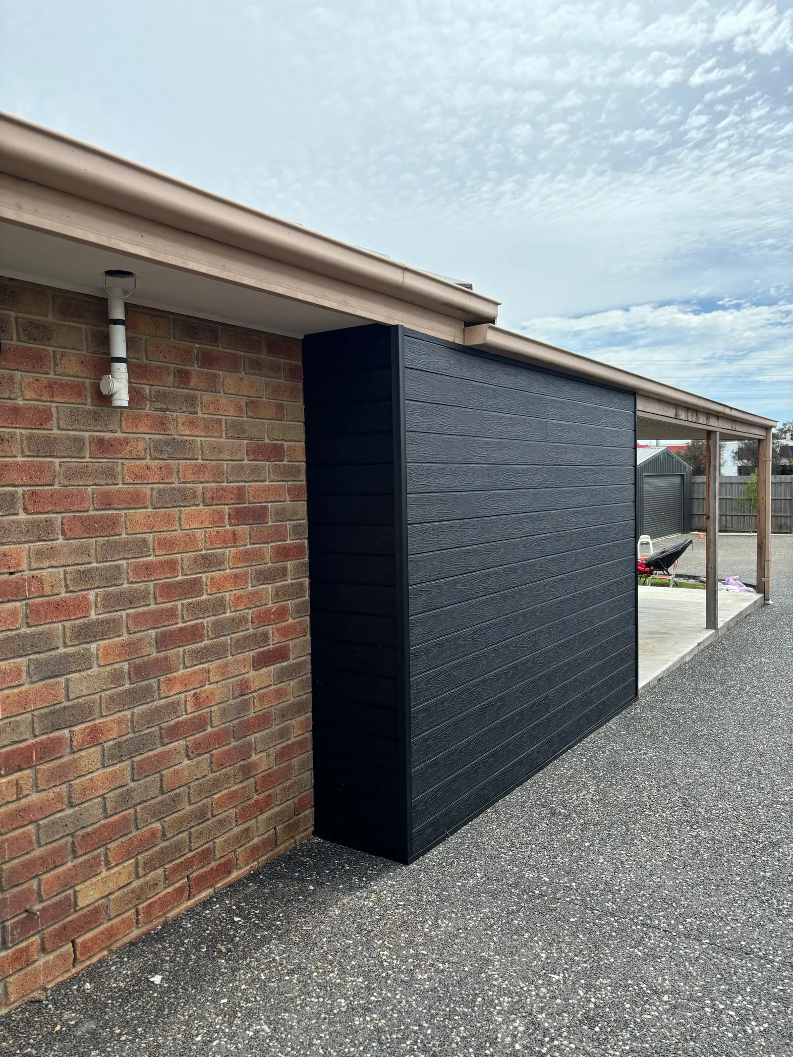 New wall clad in black woodgrain composite boards against a red brick house, with a timber-posted carport beyond