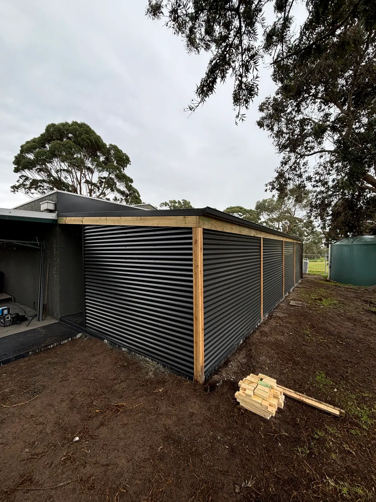 Timber-framed structure clad in dark horizontal corrugated steel with exposed timber corner post, extending from an existing building