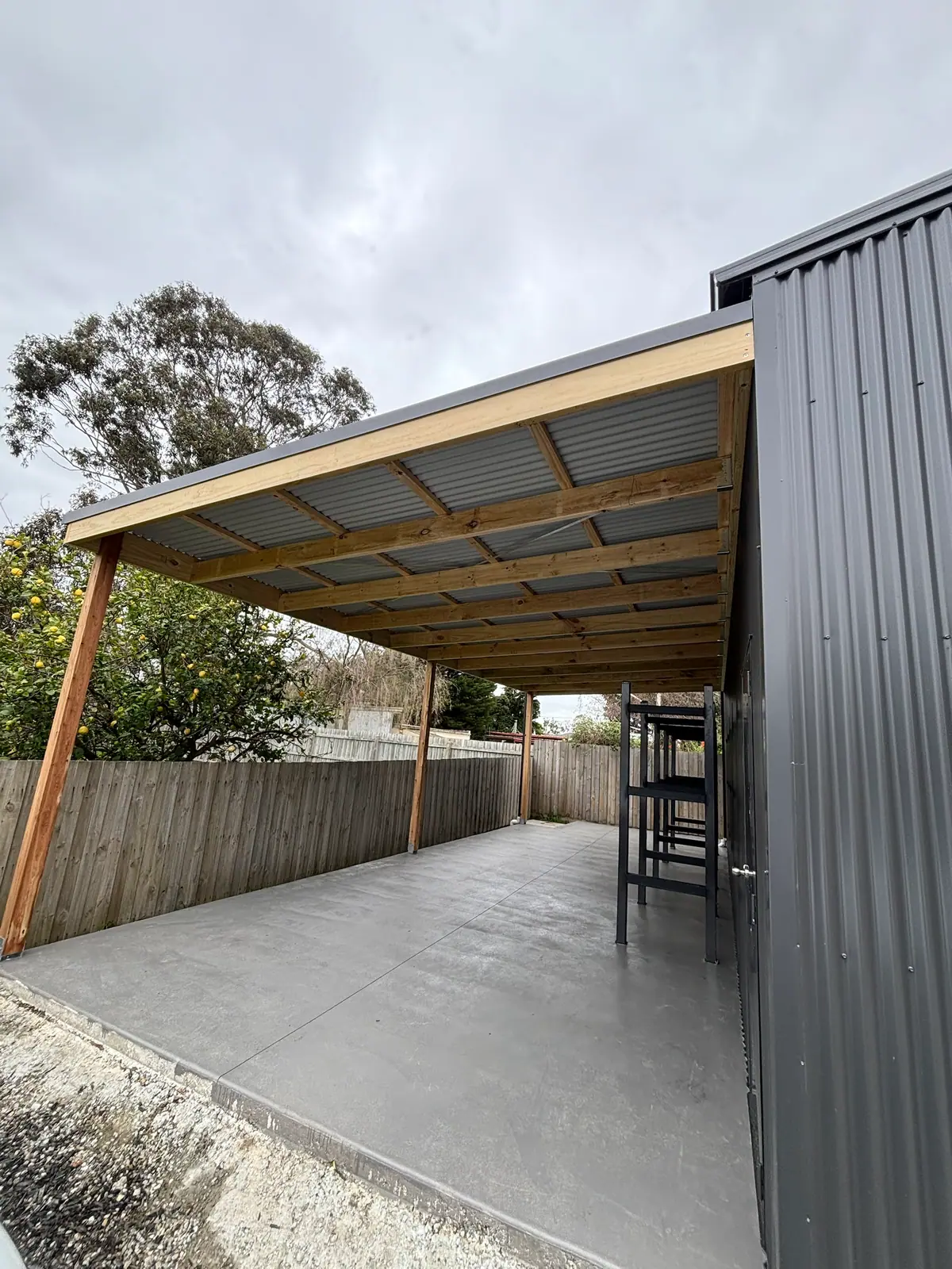 Skillion carport with exposed timber rafters and corrugated steel roof, built alongside a dark steel shed over a concrete slab
